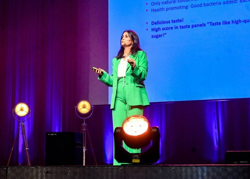 Sara Serray ler mot kameran, står framför en neutral bakgrund; klädd i professionell men avslappnad outfit, med självsäker och inspirerande hållning.